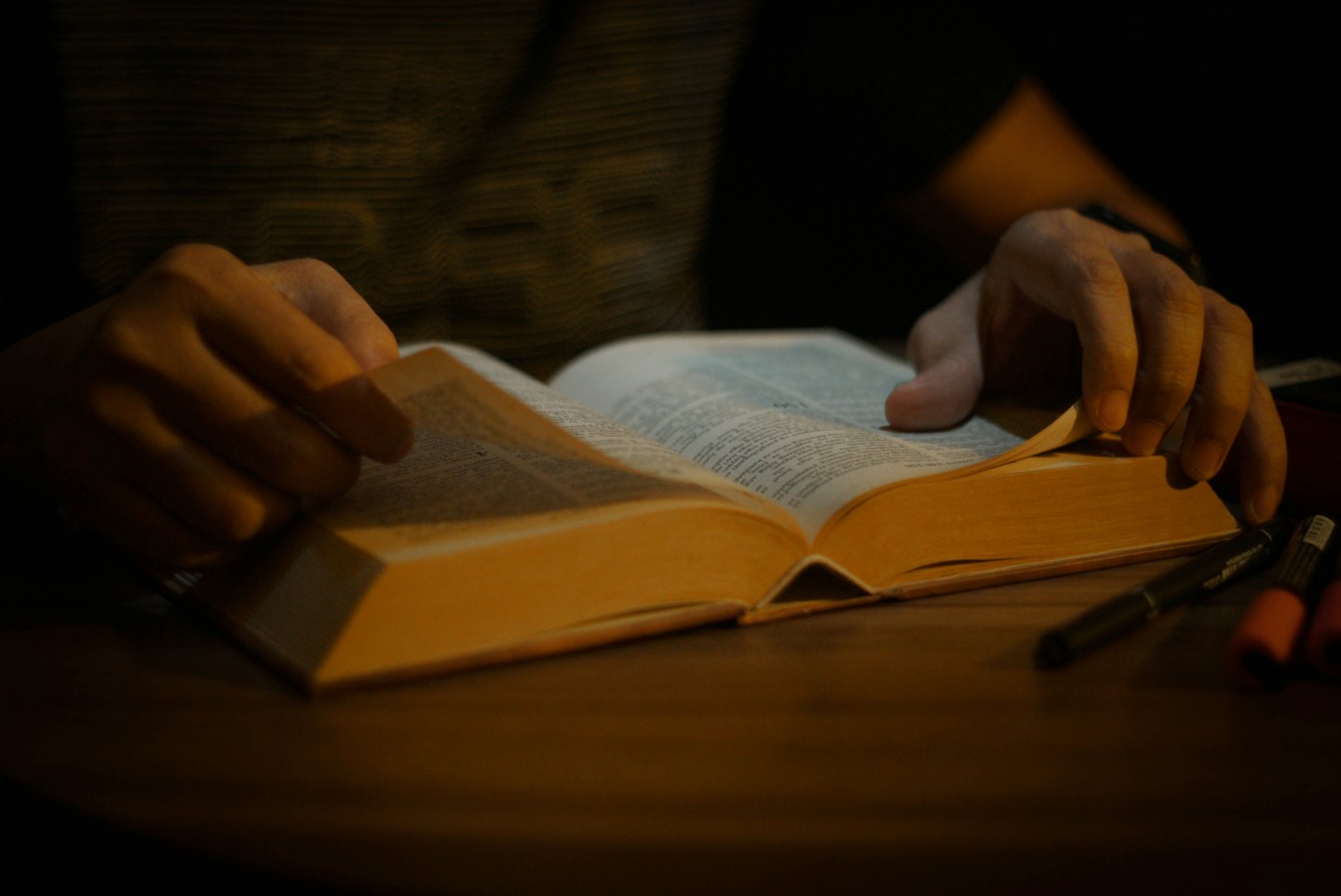 Hands turn the pages of a thick book on a wooden table.